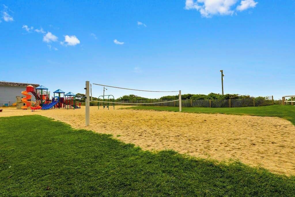 Ein Sandvolleyballfeld mit Netz, umgeben von grünem Gras und einem Spielplatz mit Rutschen.