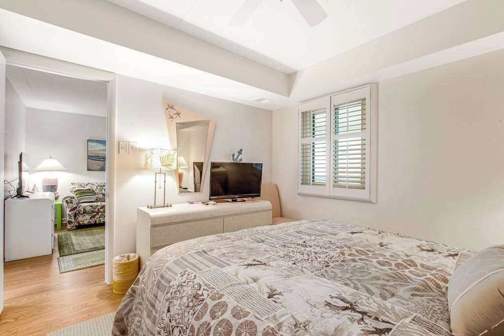 Ein Schlafzimmer mit einem Bett, einem Fernseher, einem Schreibtisch, einem Stuhl, einer Lampe und einem Fenster mit Jalousien.