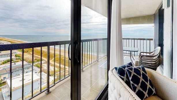 En balkon med udsigt over stranden og bygninger, et polstret siddeområde med en mønstret pude og et lille bord.