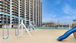 Ein Spielplatz mit Schaukeln und einer Rutsche, ein Tennisplatz und im Hintergrund ein Hochhaus.