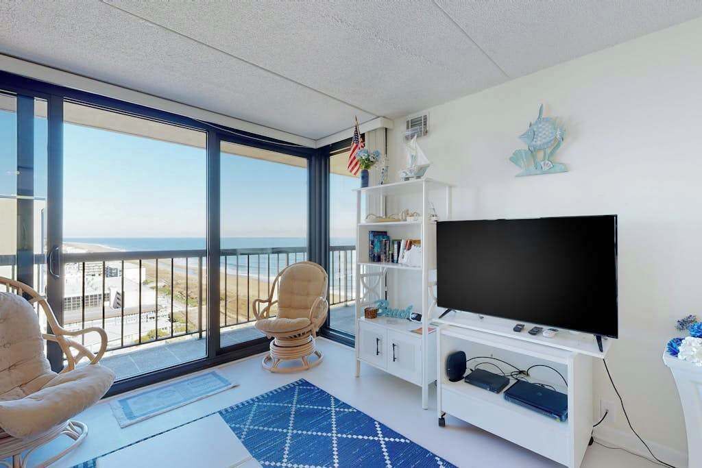 Ein Zimmer mit großem Fernseher, einer weißen Unterhaltungsanlage, einem Korbstuhl und Blick auf den Strand.