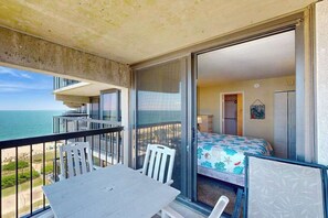 A balcony with a table and chairs, a bed with a floral bedspread, and a view of the ocean. - Beach condo with balcony, W/D, pools, game room, tennis & gym (Ocean City)
