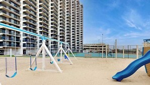 A playground with swings and a slide, a tennis court, and a high-rise building in the background. - Bright, ocean-view condo with balcony, pools, tennis, saunas & gym (Ocean City)