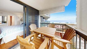 A balcony with a wooden table and chairs, a bed with a patterned bedspread, and a view of the ocean. - Ocean-view condo with balcony, AC, pools, saunas, tennis & gym (Ocean City)