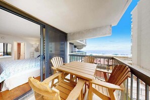 A balcony with a wooden table and chairs, a bed with a patterned bedspread, and a view of the ocean. - Ocean-view condo with balcony, AC, pools, saunas, tennis & gym (Ocean City)
