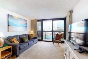 A living room with a sofa, armchair, TV, and a balcony with a view. - Convenient & breezy condo with balcony, pools, tennis, sauna & gym (Ocean City)