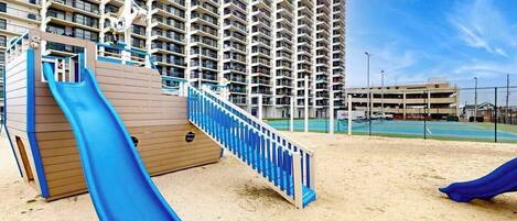 A playground with a blue slide and a wooden structure, set against a backdrop of a tall residential building.