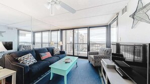 A modern living room with a sofa, coffee table, TV, and large windows offering a city view. - Ocean-view getaway with balcony, AC, pools, saunas, tennis & gym (Ocean City)