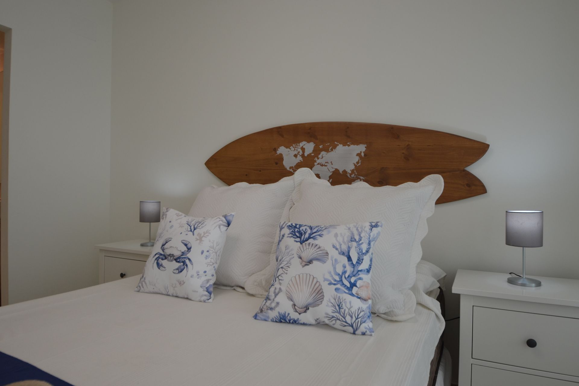 Un lit avec une literie blanche et deux coussins décoratifs, une tête de lit en bois et deux lampes de chevet posées sur des tables de chevet blanches.