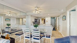 A modern living room with a glass dining table, white chairs, a blue sofa, and a ceiling fan. - Sunny coastal condo with beach views, balcony, pools, gym, sauna & AC (Ocean City)