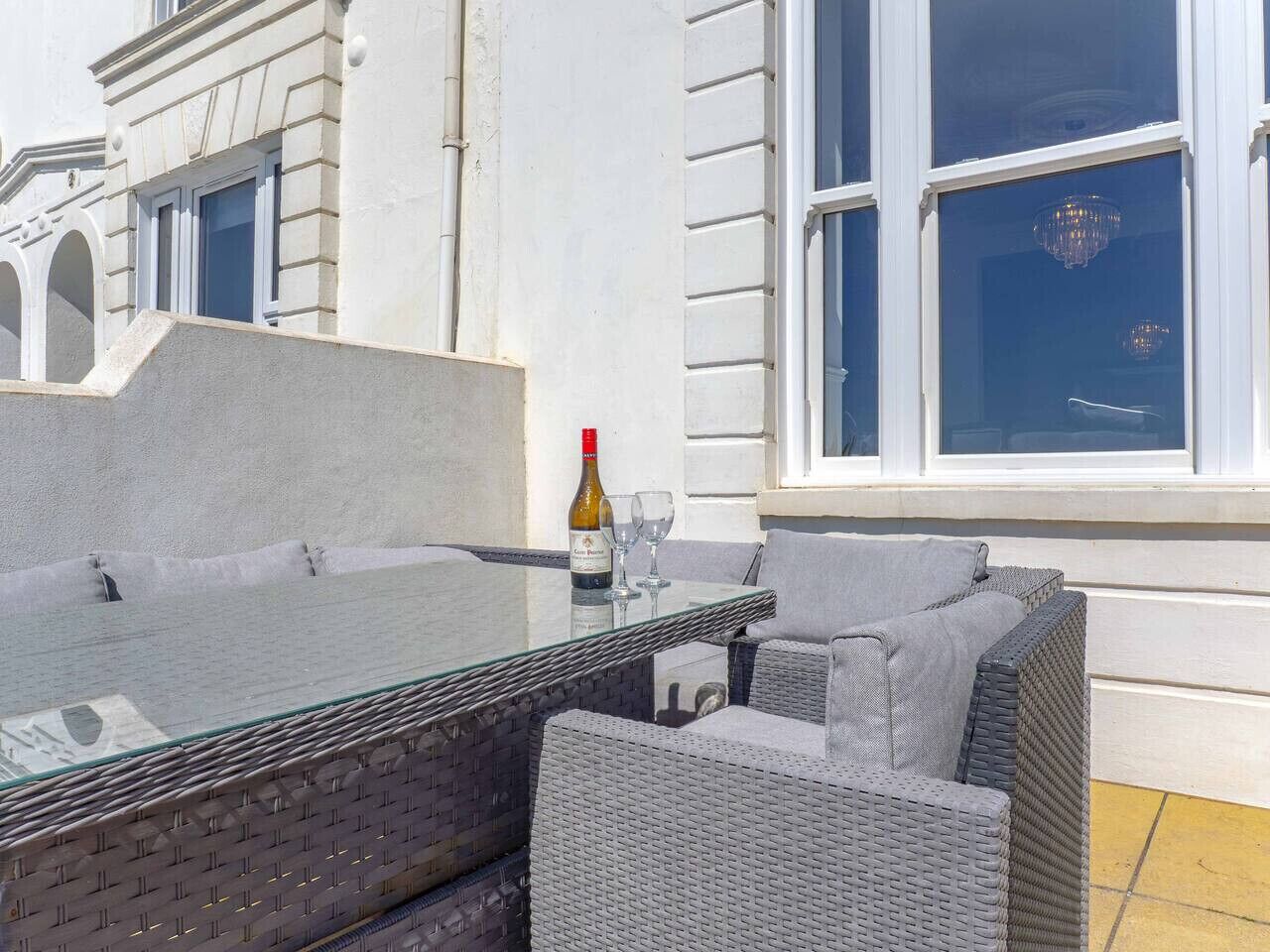 Een tafel met glazen blad, omringd door rieten stoelen, een fles wijn en wijnglazen.