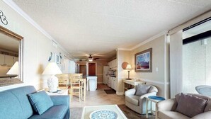 A living room with a sofa, a coffee table, a dining area with chairs, and a kitchen in the background. - Updated, oceanfront condo with furnished balcony, fast WiFi, central AC & pool (Ocean City)