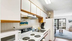 Cozinha compacta com micro-ondas, fogão e pia, armários de madeira e vista para uma varanda.