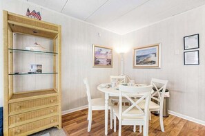 Ein Essbereich mit einem runden Tisch und weißen Stühlen, eine Korbmöbel-Anrichte mit Glastüren sowie gerahmte Bilder an der Wand.