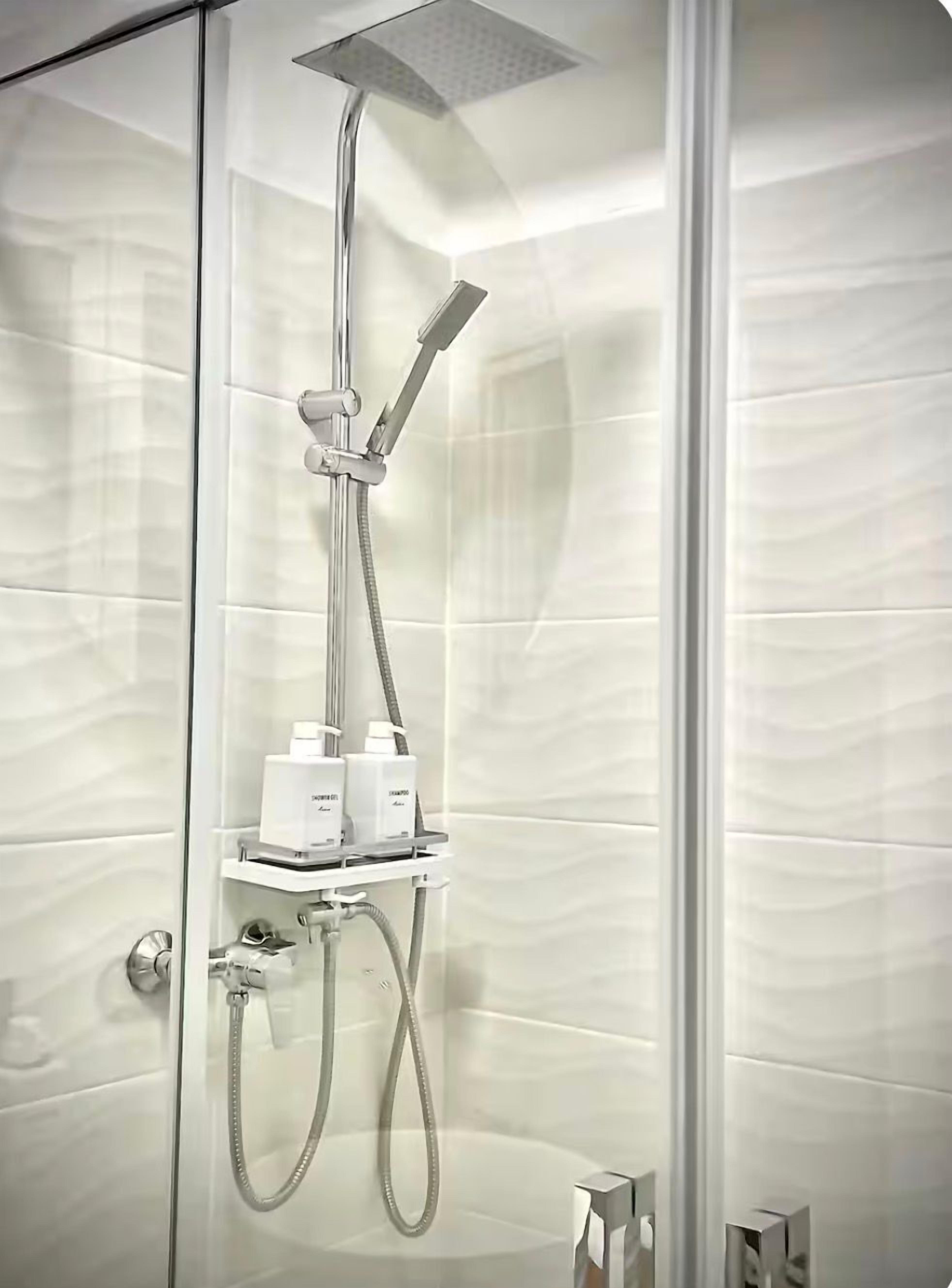 Une douche fermée par des parois en verre, dotée d’un pommeau de douche à main et d’un pommeau de douche fixe au plafond, d’un panneau de commande et d’une étagère contenant deux bouteilles.