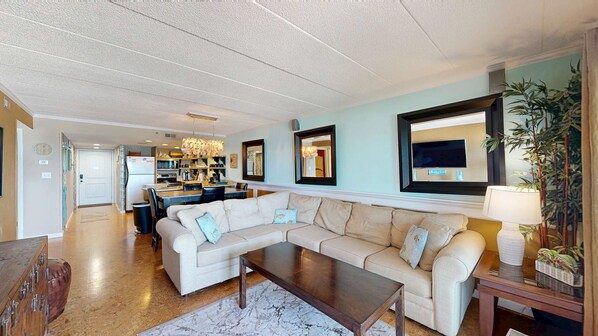 A living room with a sectional sofa, a coffee table, and a TV mounted on the wall. - Sandpiper Dunes 504 (Ocean City)