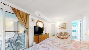 A hotel room with a bed, a television on a wooden dresser, a chair, a glass door leading to a balcony, and a painting on the wall. - Sandpiper Dunes 212 (Ocean City)
