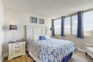 A bedroom with a bed, a nightstand, a lamp, a window with curtains, and a view of buildings. - Sunny condo with balcony, W/D, pools, tennis & AC (Ocean City)