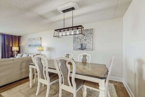 A dining area with a wooden table and white chairs, a chandelier, and a painting on the wall. - Sunny condo with balcony, W/D, pools, tennis & AC (Ocean City)