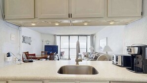 A kitchen with a large sink, a dining area with chairs, a sofa, and a coffee maker. - Oceanfront Condo with a Shaded Balcony, AC, WiFi, Laundry & Shared Pool (Ocean City)