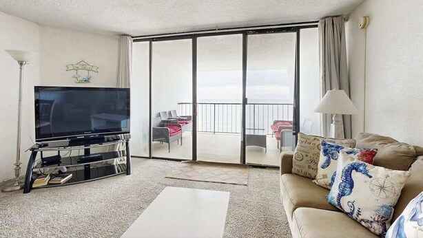 A living room with a flat-screen TV on a glass-top table, a sofa with decorative pillows, a lamp, and a sliding glass door leading to an outdoor seating area. - Oceanfront Condo with a Shaded Balcony, AC, WiFi, Laundry & Shared Pool (Ocean City)
