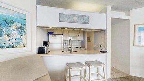 A compact kitchen with a microwave, coffee maker, and a sink. There are two stools and a couch in the living area. - Oceanfront Condo with a Shaded Balcony, AC, WiFi, Laundry & Shared Pool (Ocean City)