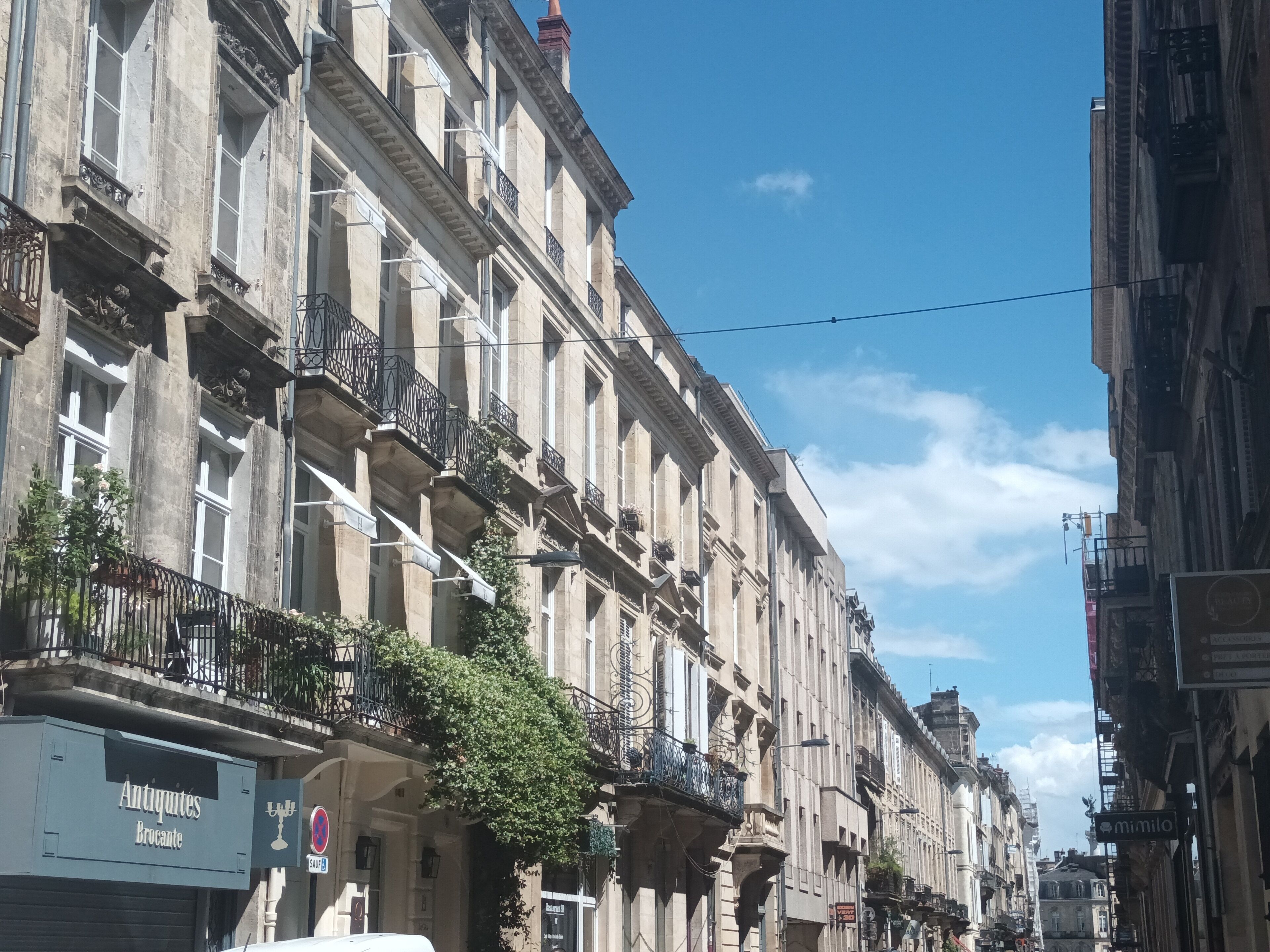 A row of historic buildings with balconies, a sign for 'Antiquités Brocante,' and a 'mimilio' sign.