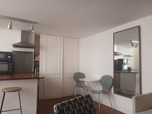 Eine kompakte Küche mit Mikrowelle, Herd und Deckenleuchten. Zwei Stühle und ein kleiner Tisch stehen bereit, an der Wand befindet sich ein großer Spiegel.