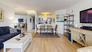 Ein modernes Wohnzimmer mit einer Couch, Couchtisch und Fernseher. Es gibt einen Essbereich mit Tisch und Stühlen, und eine Küche im Hintergrund.