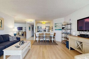 A modern living room with a sofa, coffee table, and TV. There is a dining area with a table and chairs, and a kitchen in the background. - Up-to-Date, Oceanfront Condo with Balcony, AC, WiFi, Laundry & Shared Pool (Ocean City)