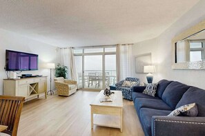 A living room with a sofa, a coffee table, a TV, and a balcony with a view. - Up-to-Date, Oceanfront Condo with Balcony, AC, WiFi, Laundry & Shared Pool (Ocean City)