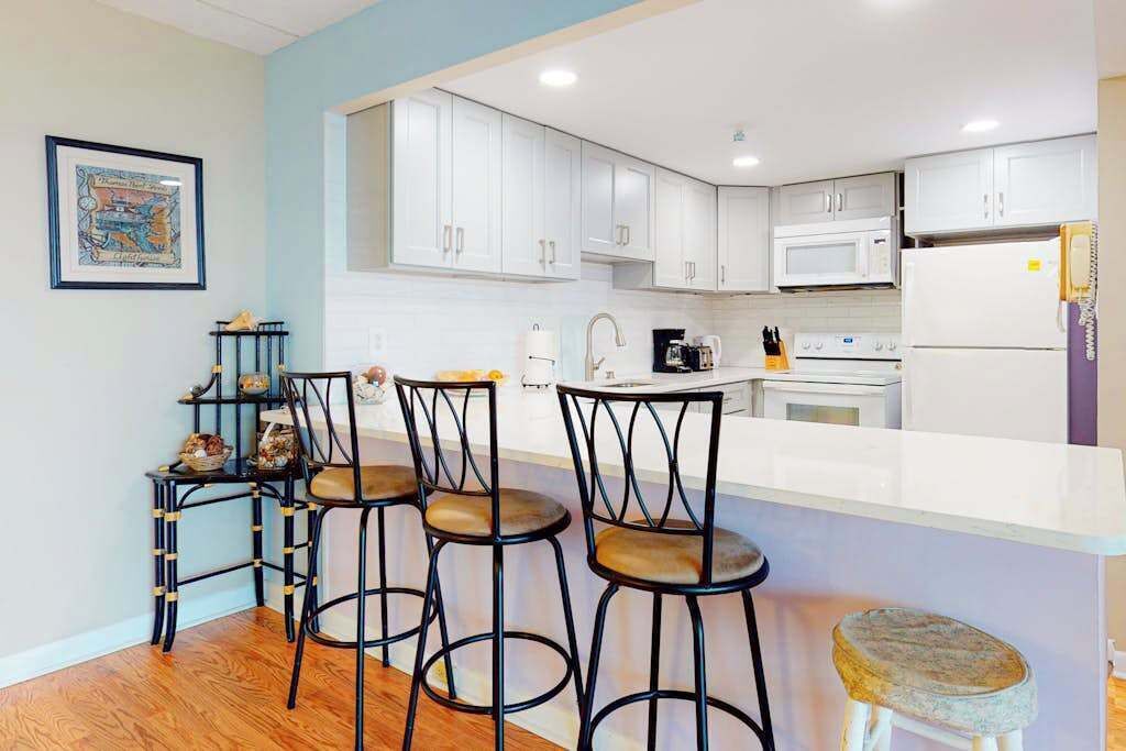 Uma cozinha moderna com armários brancos, bancada clara e um espaço bar com banquetas.