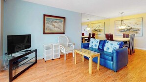 Ein Wohnzimmer mit Fernseher, weißem Sessel, blauem Sofa, Couchtisch aus Holz und einem Essbereich mit Stühlen und Tisch.