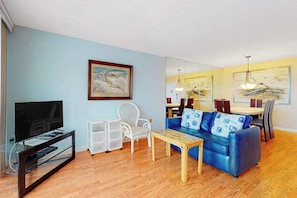 A living room with a TV, white chair, blue sofa, wooden coffee table, and a dining area with chairs and a table. - Convenient condo with side ocean views, balcony, pools, & tennis (Ocean City)