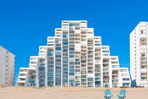 A multi-story hotel with balconies, a sandy ground floor, and a clear blue sky. - Oceanfront Condo with Beach Views, Balcony, AC, WiFi & Shared Outdoor Pool (Ocean City)