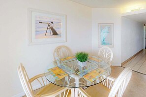 A dining area with a glass table, white chairs, and framed artwork on the walls. - Oceanfront Condo with Beach Views, Balcony, AC, WiFi & Shared Outdoor Pool (Ocean City)