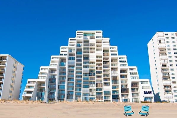 A modern multi-story hotel with balconies, a sandy foreground, and clear blue sky. - Oceanfront Condo with a Balcony, Beach Views, AC, WiFi & Shared Outdoor Pool (Ocean City)