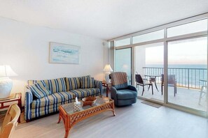 A living room with a sofa, coffee table, and a chair, a balcony with a view of the sea, and a table with chairs. - Oceanfront Condo with a Balcony, Beach Views, AC, WiFi & Shared Outdoor Pool (Ocean City)