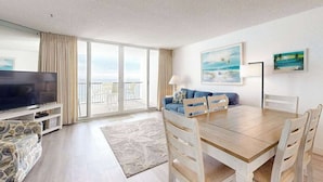 Ett rymligt vardagsrum med en platt-TV, ett matbord med stolar, en soffa och en balkong med utsikt.
