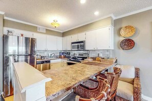 A kitchen with white cabinets, stainless steel appliances, a granite countertop, wicker chairs, and a ceiling light fixture. - Our Place At The Beach 206B (Ocean City)
