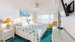 Une chambre à coucher comprenant un lit, un ventilateur de plafond, une télévision, une fenêtre avec des stores et une chaise.