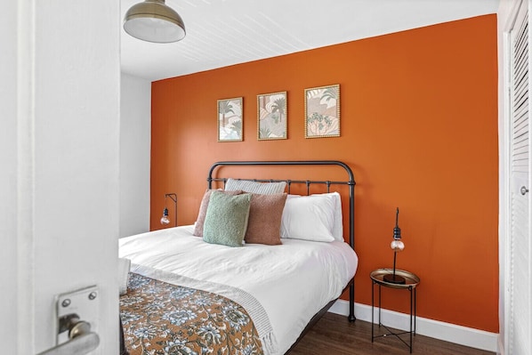 Une chambre avec un mur orange, un lit avec une literie blanche et trois tableaux encadrés au-dessus du lit.
