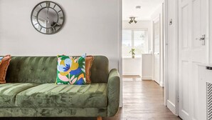 A green sofa with a colorful pillow, a wall clock, and a white door in a room with wooden flooring. - Modern 2-Bed Apartment in Reading (Berkshire)