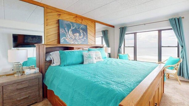 Ein Schlafzimmer mit einem großen Bett, einem hölzernen Kopfteil, einem Gemälde, einem Nachttisch, einem Stuhl und einem Fenster mit Vorhängen.