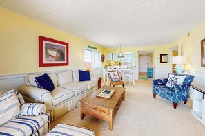 Ein Wohnzimmer mit einer Couch, einem Couchtisch und einem gemusterten Sessel. Im Hintergrund befindet sich ein Essbereich mit Tisch und Stühlen.