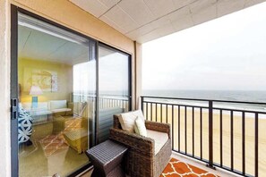Un balcon avec une chaise en osier et une petite table, une porte vitrée donnant sur une pièce, et une vue sur la plage.