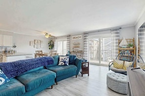 A living room with a sectional sofa, a round ottoman, a bookshelf, and a dining area with a table and chairs. - Second-Floor Condo 1 Block from the Beach w/ Balcony, AC, WiFi & Shared Pool (Ocean City)