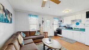 Ein Wohnzimmer mit braunem Sofa, einem Couchtisch und einer Küchenzeile mit weißen Schränken und einer Mikrowelle.