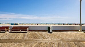 Eine Holzterrasse mit Bänken und einem Mülleimer, mit Blick auf einen Strand mit Sonnenschirmen und Menschen.