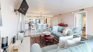 Uma sala de estar moderna com sofá, mesa de centro, TV de tela plana na parede e uma área de jantar ao fundo.